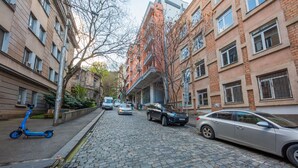 Una calle adoquinada flanqueada por edificios de varios pisos, coches aparcados y un patinete azul.