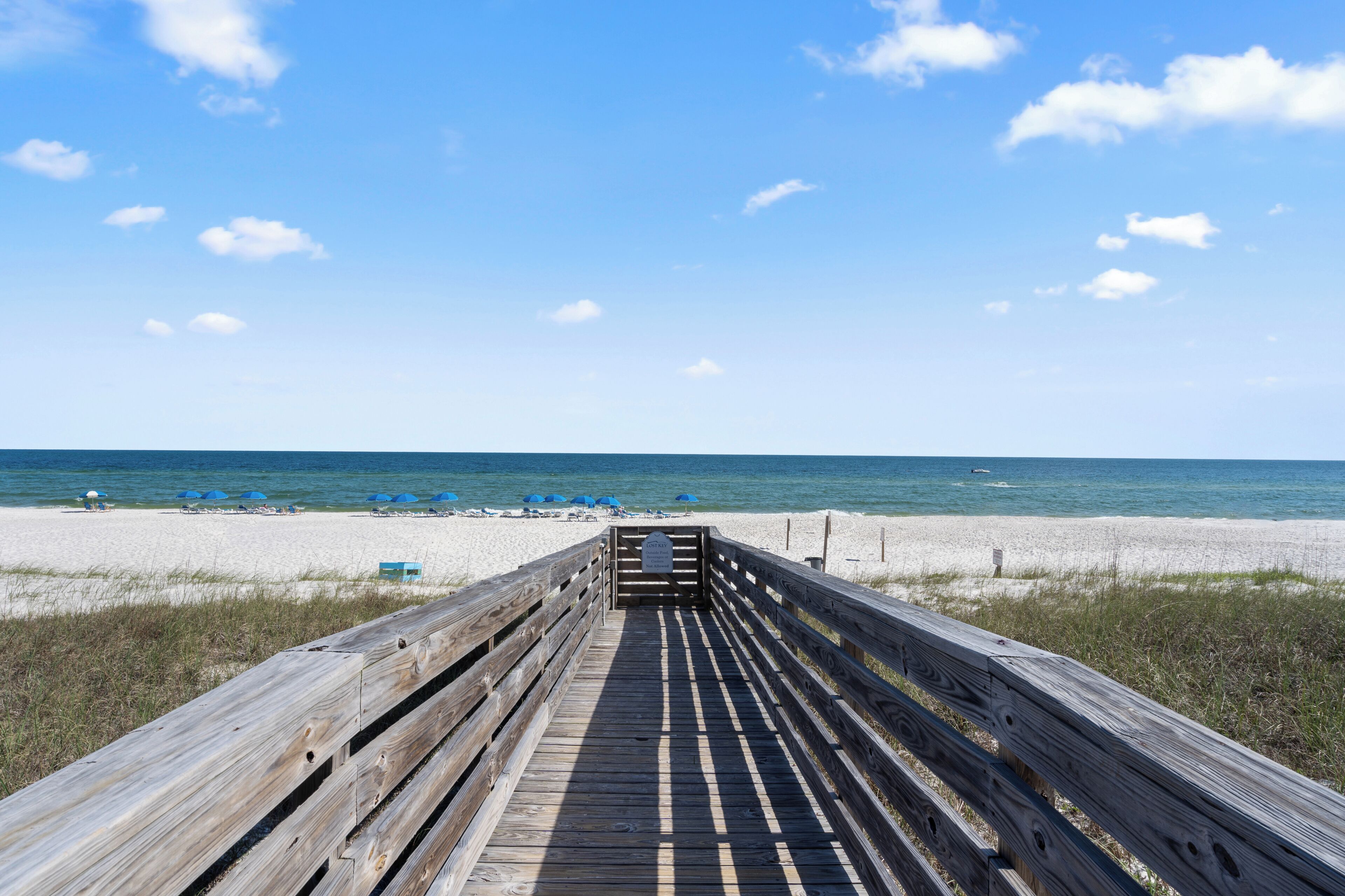 A wooden boardwalk leading to a beach with blue umbrellas and lounge chairs.