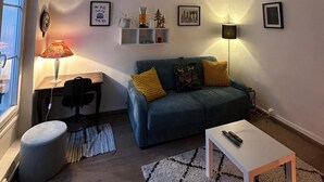 Una sala de estar con un sofá, una mesa, una lámpara, un estante pequeño con objetos decorativos y un ventanal.