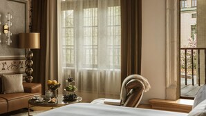 Habitación de hotel con cama, sofá, mesa con frutas y tetera, lámpara y vista a un balcón.