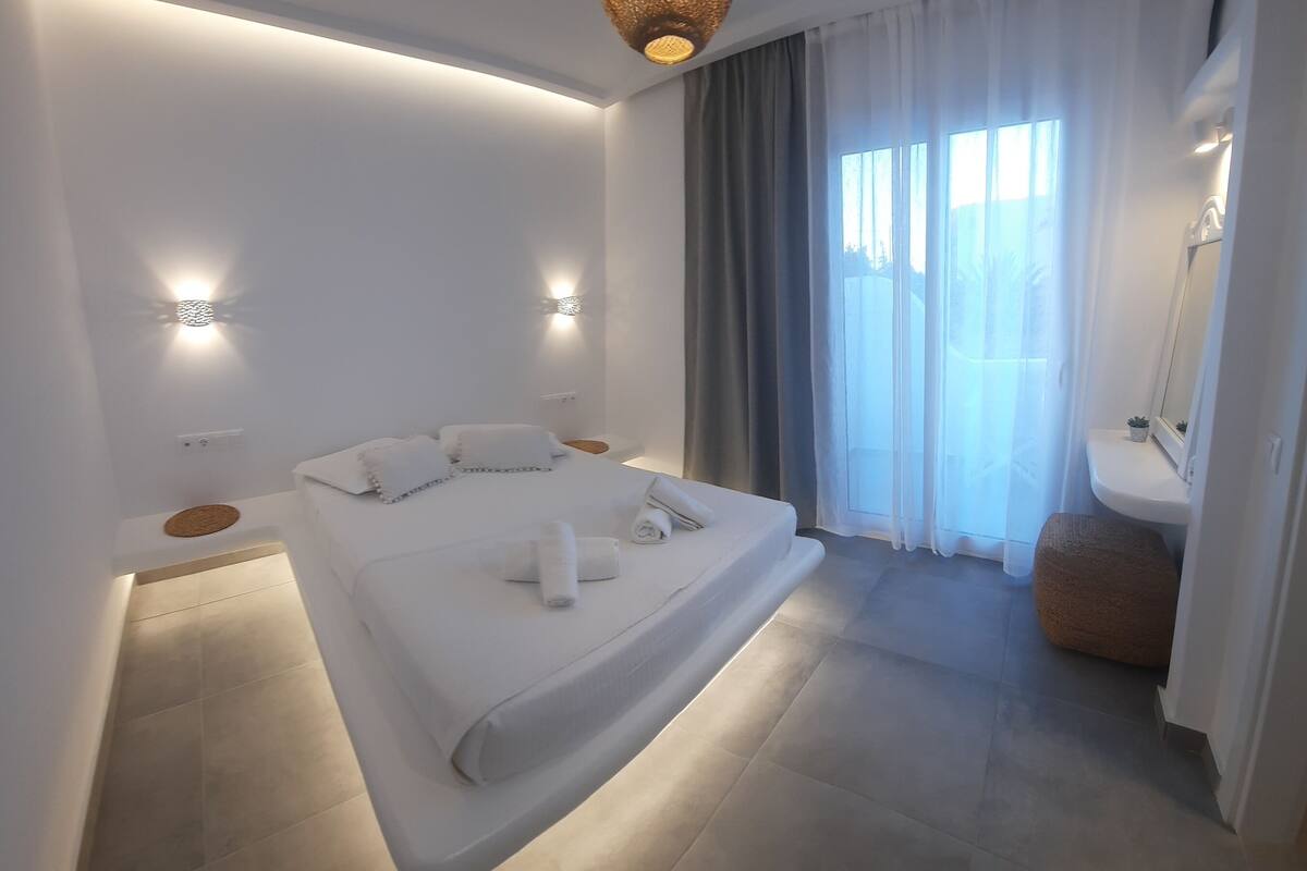 Un dormitorio moderno con cama, mesitas de noche, lámpara colgante, ventana con cortinas y baño con lavabo y espejo.