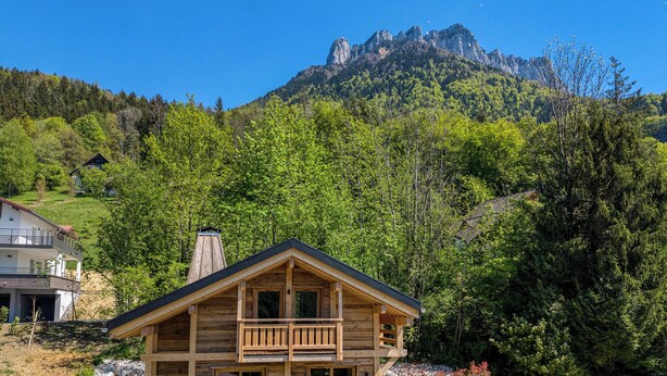 Eine Holzhütte mit Balkon, umgeben von üppigem Grün und einer Bergkulisse.