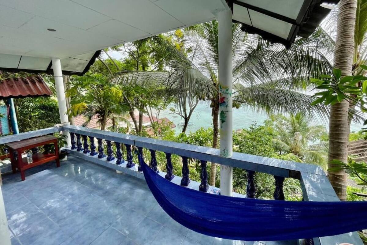 Ein überdachter Balkon mit einer blauen Hängematte, Palmen und Blick aufs Meer.