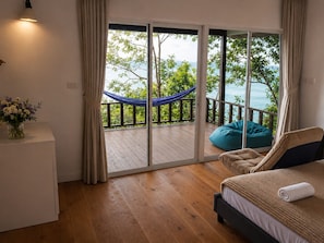 Un dormitorio con suelo de madera, cama con cabecera beige y oscura, una hamaca azul en un balcón y vistas a la vegetación y al agua.