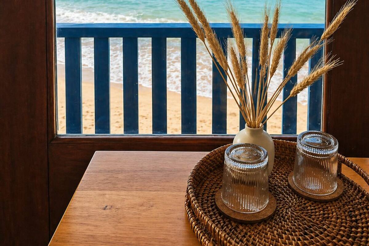 Blick auf das Meer durch ein Fenster, mit einem Holztisch, einer Vase mit Weidenähren und zwei Glaswindlichtern.