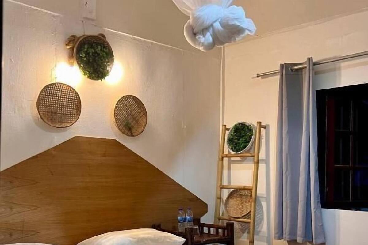 Ein ordentlich bezogenes Bett mit Kissen und einem gefalteten Handtuch, ein hölzernes Kopfende, eine Wandleuchte und ein Fenster mit Vorhängen.