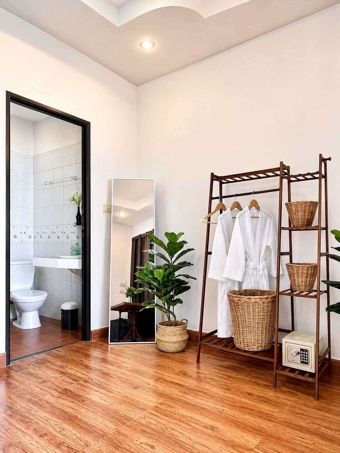 Een moderne badkamer met een houten vloer, een wit toilet, een wastafel, een spiegel en een houten rek met witte gewaden en manden.