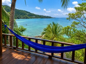 A wooden deck with a blue hammock, overlooking a turquoise sea and a green hillside.