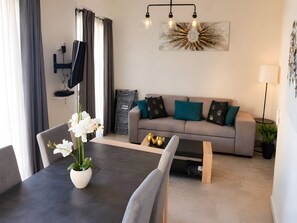 Ein modernes Wohnzimmer mit einer Couch, einem Couchtisch und einem Esstisch mit Stühlen. An der Wand ist ein Fernseher angebracht, es gibt eine Pflanze und ein dekoratives Wandbild.