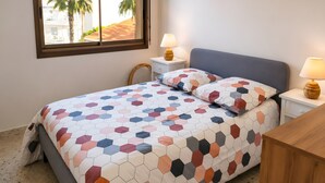 Een slaapkamer met een bed, twee nachtkastjes, een raam met uitzicht op palmbomen en een lamp op elk nachtkastje.