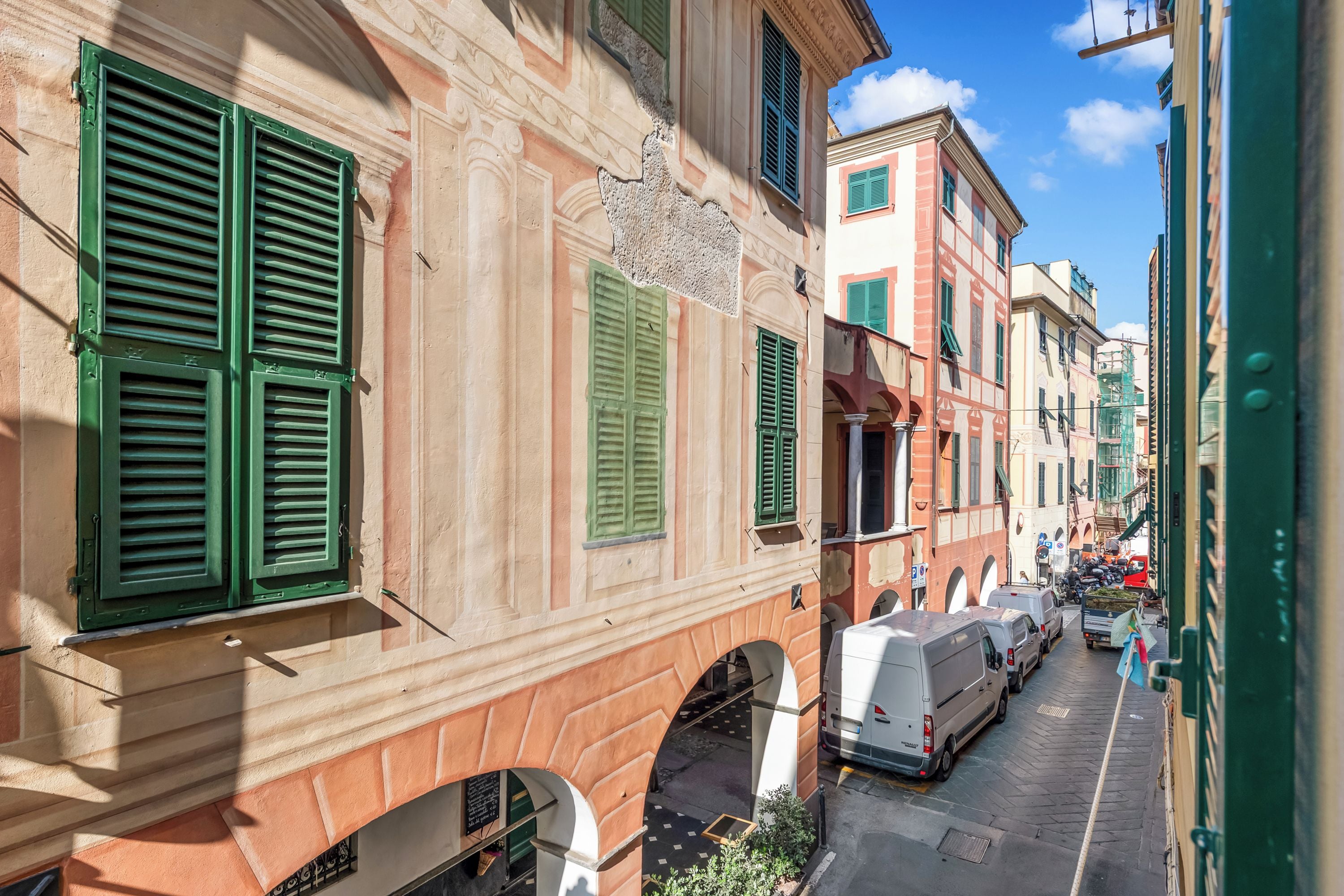 Una calle estrecha con edificios históricos, contraventanas verdes y vehículos estacionados.