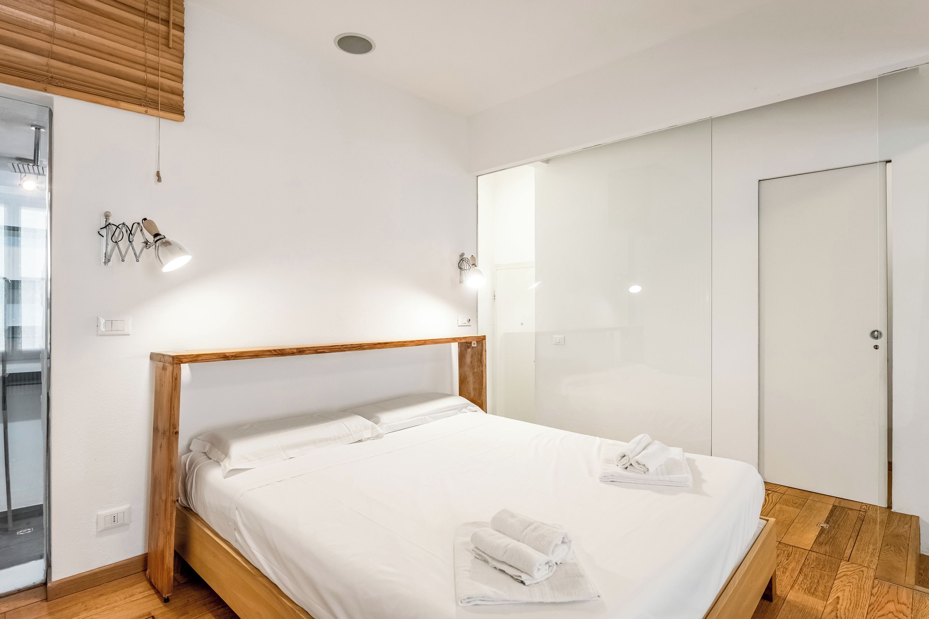 A modern bedroom with a wooden bed frame, white linens, a glass shower enclosure, and a white wardrobe.