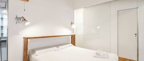 A modern bedroom with a wooden bed frame, white linens, a glass shower enclosure, and a white wardrobe.