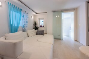 Ein modernes Wohnzimmer mit einem weißen Sofa, einer Glastür, die auf einen Balkon führt, und einem kleinen Fenster mit einer Pflanze.