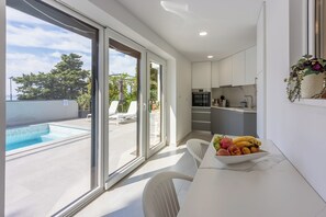 Eine moderne Küche mit weißen Schränken, ein Essbereich mit Obstschale und Blick zum Pool.