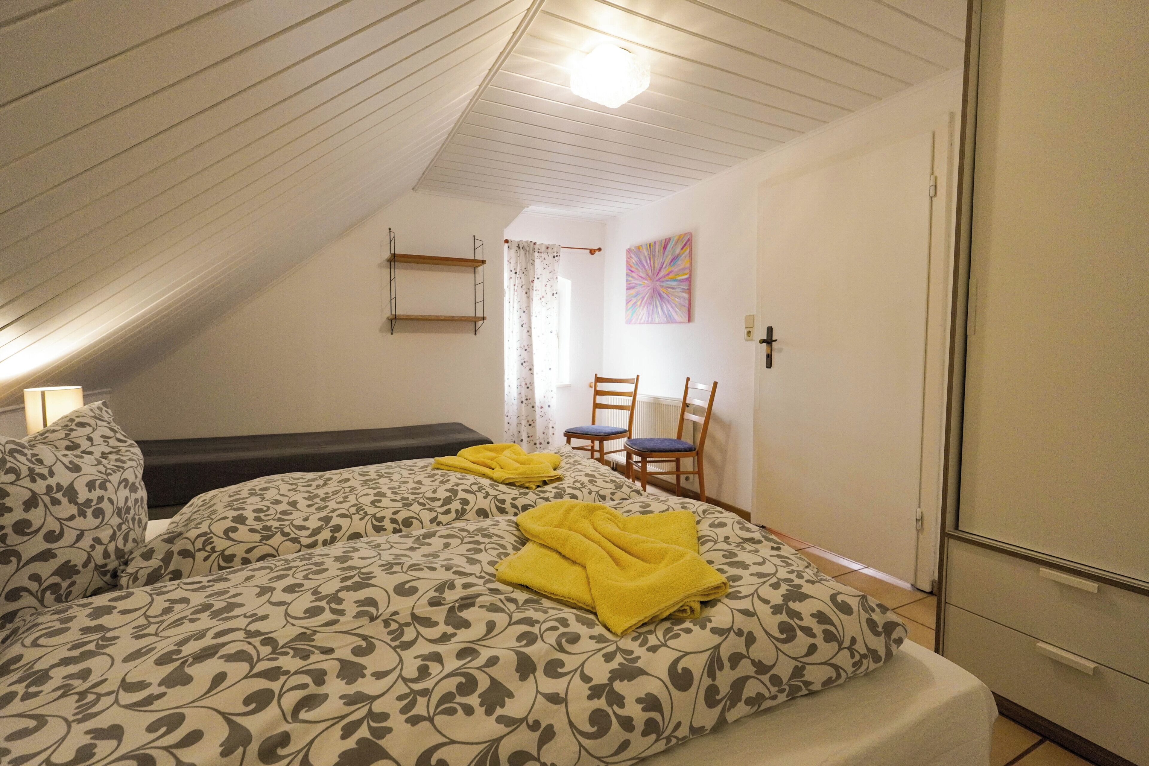 A room with a sloped ceiling, a bed with patterned bedding, a chair, a desk, and a radiator.