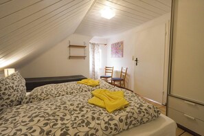 A room with a sloped ceiling, a bed with patterned bedding, a chair, a desk, and a radiator.