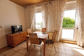 A dining area with a table, chairs, a television on a wooden cabinet, and large windows with floral curtains.