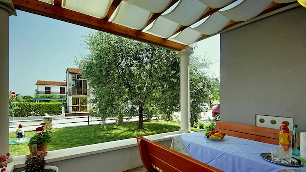 Ein Balkon mit einer Holzbank und einem Tisch, Blumentöpfen und Blick auf ein Gebäude und Grünflächen.