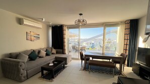 A modern living room with a sofa, coffee table, dining table, and chairs. There is a TV, a ceiling light, and a large window with curtains. - Fouka North Coast CL3-CH19-02 (Marsa Matrouh)