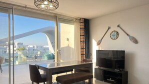 A modern living room with a glass sliding door leading to a balcony, a flat-screen TV, and a dining area with a bench. - Fouka North Coast CL3-CH19-02 (Marsa Matrouh)