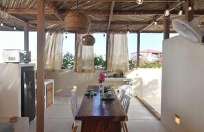 A covered outdoor dining area with a table set for two, a fan, and a refrigerator in the background. - Loft Boutique *Starlink | Private Pool * La Punta (La Barra de Colotepec)