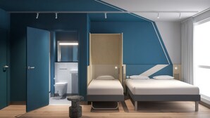 A modern bedroom with two beds, a small seating area, and a bathroom visible through an open door. - Ibis Budget Guingamp - Coeur De Bretagne (Ouverture Septembre 2026) (Ploumagoar)