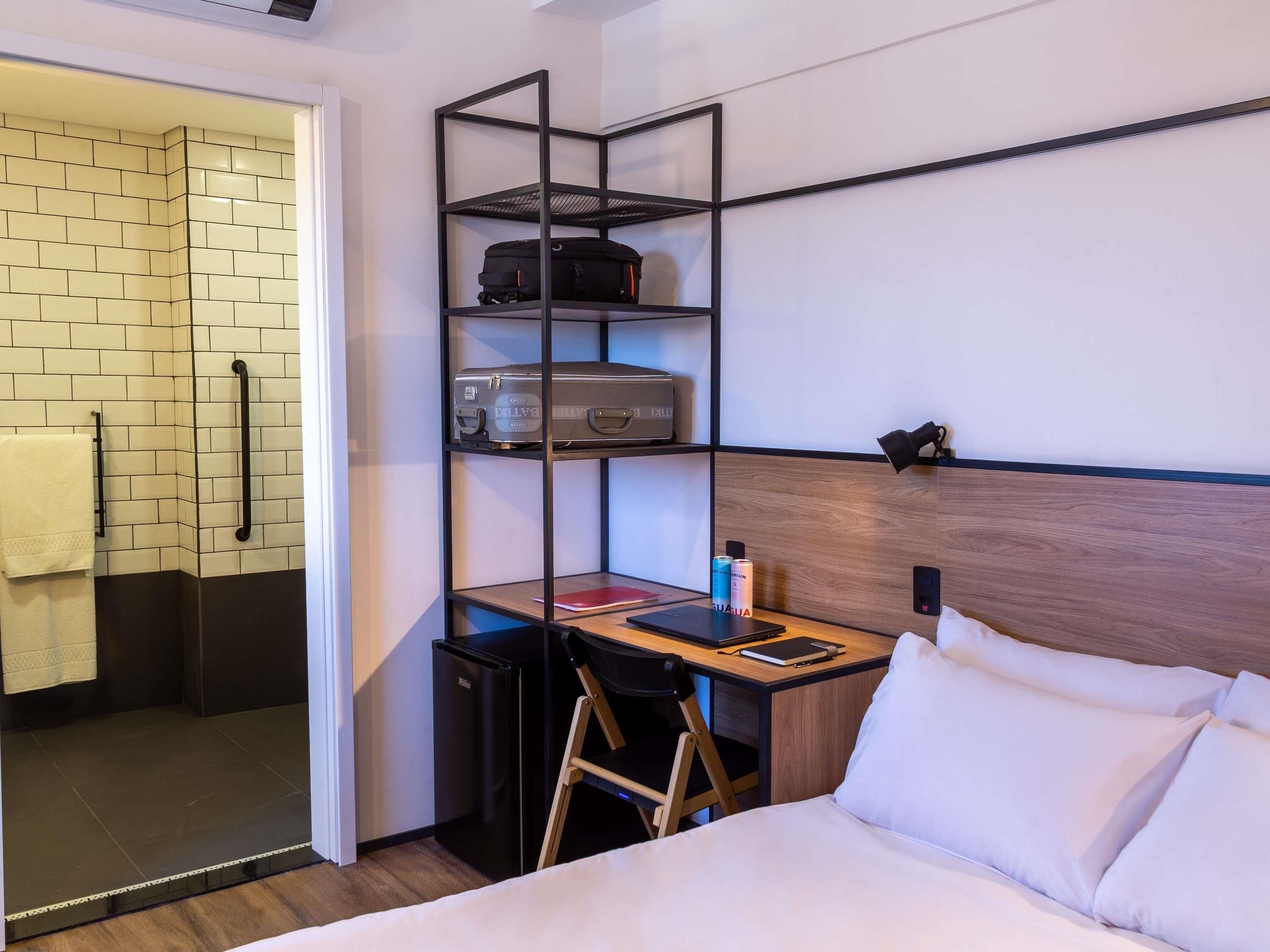 A hotel room with a bed, a desk with a chair, a small kitchenette, a bathroom with a towel rack, and a shelf with luggage.