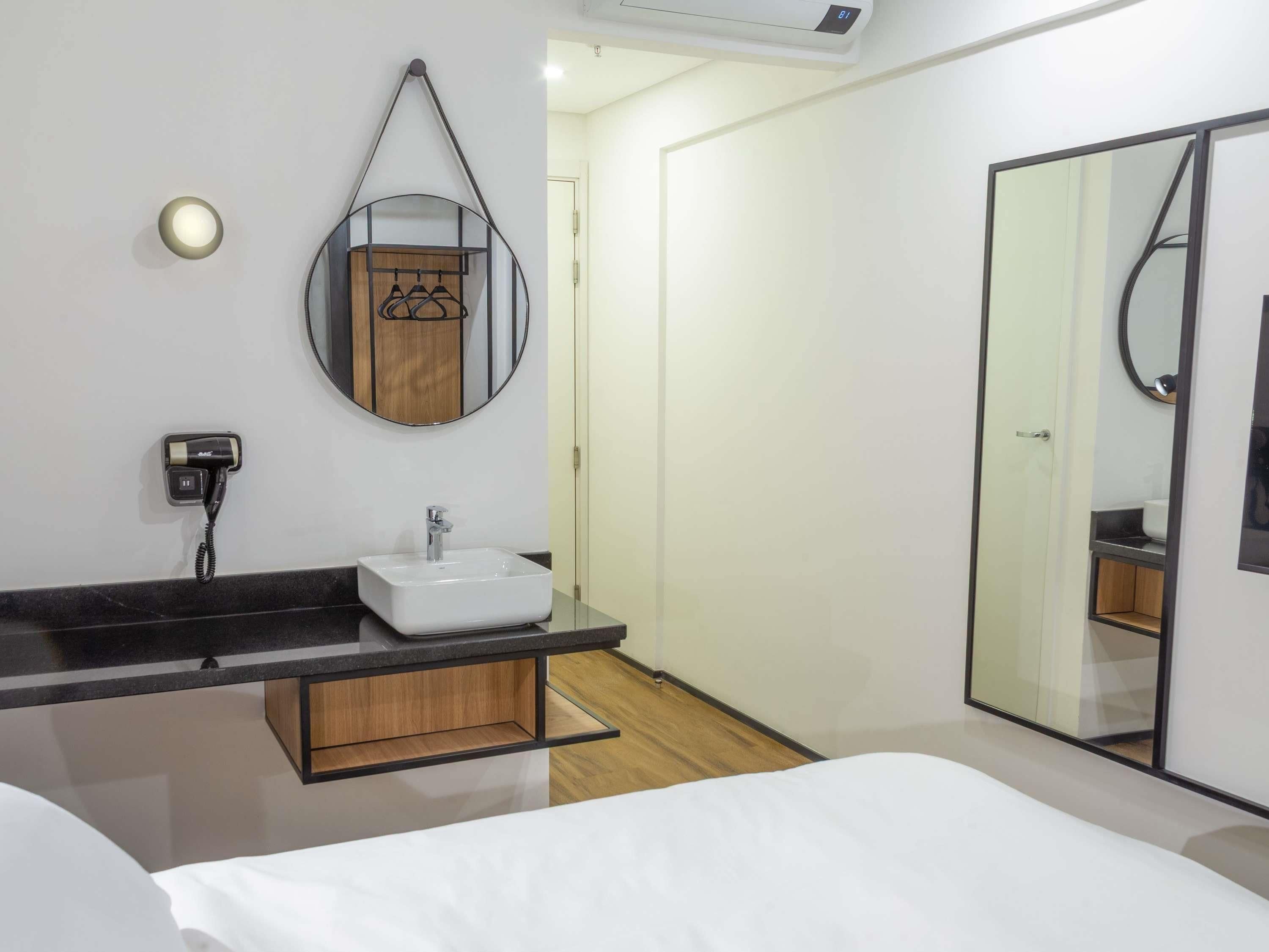 Ein modernes Hotelzimmer mit einem großen Bett, einem Nachttisch, einem Badezimmer mit Waschbecken und Spiegel sowie einem Kleiderschrank.