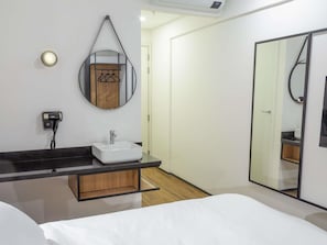 Una habitación de hotel moderna con una cama grande, una mesita de noche, un baño con lavabo y espejo, y un armario.
