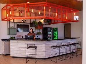 Una zona de bar con techo de malla roja, un bartender detrás de la barra y un letrero de neón verde.