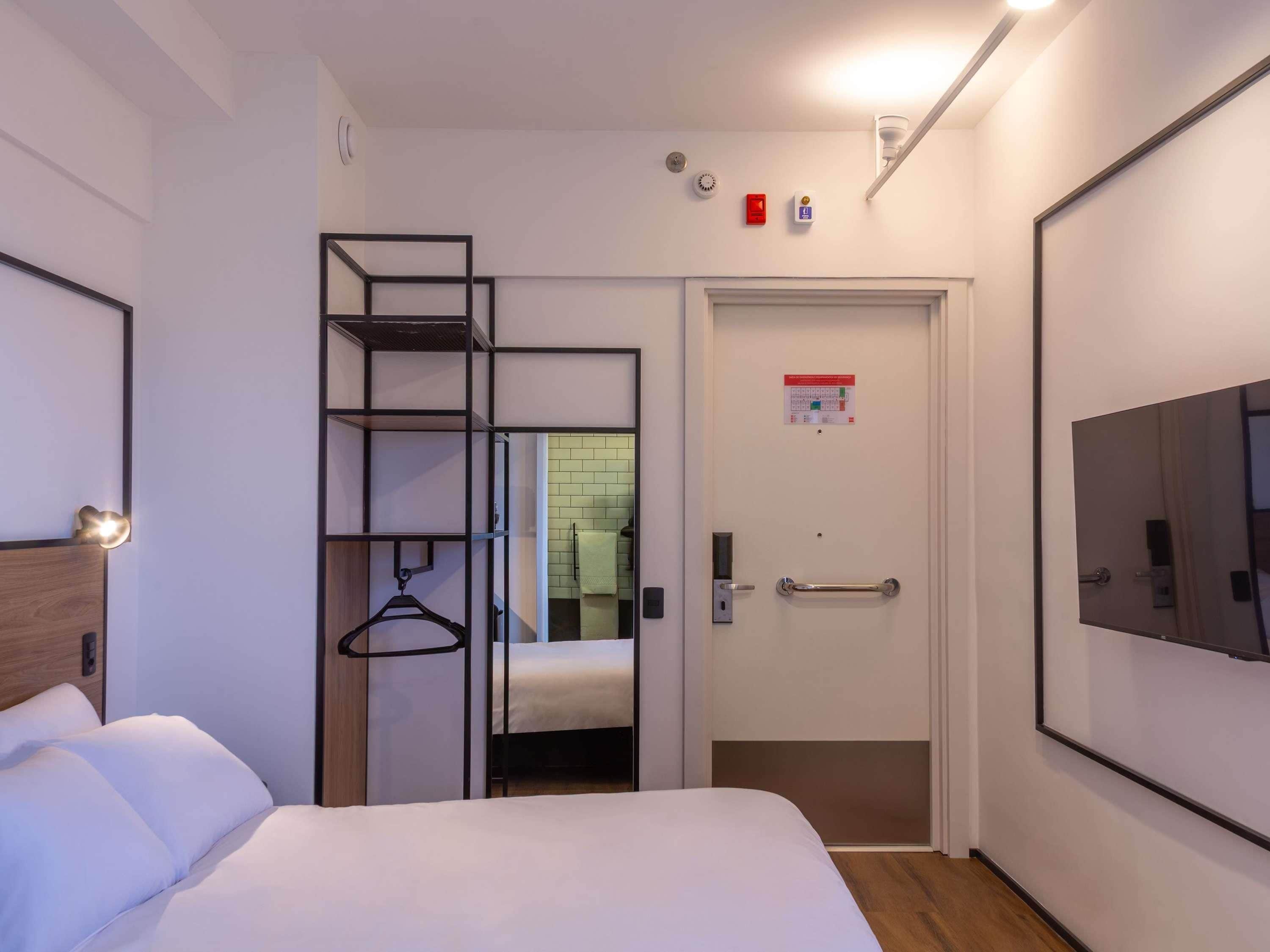 Quarto de hotel com cama, guarda-roupa com espelho, porta com puxador e televisão presa à parede.