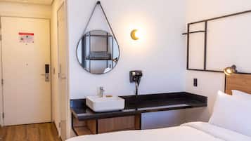 Habitación de hotel con cama, mesita de noche, espejo, lámpara de pared y estante con una maleta.