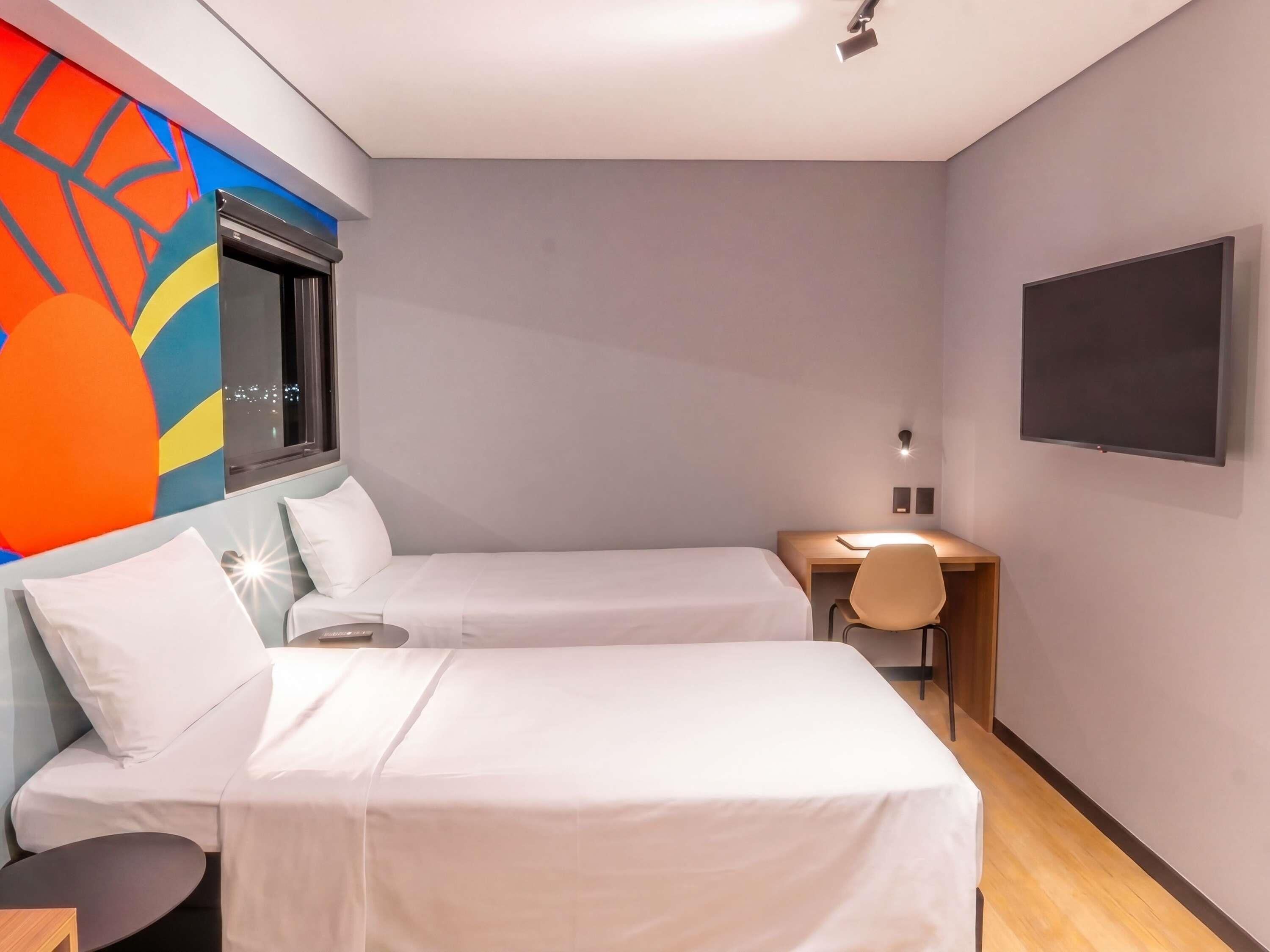 A hotel room with two beds, a desk, a chair, a TV, and a colorful wall mural.