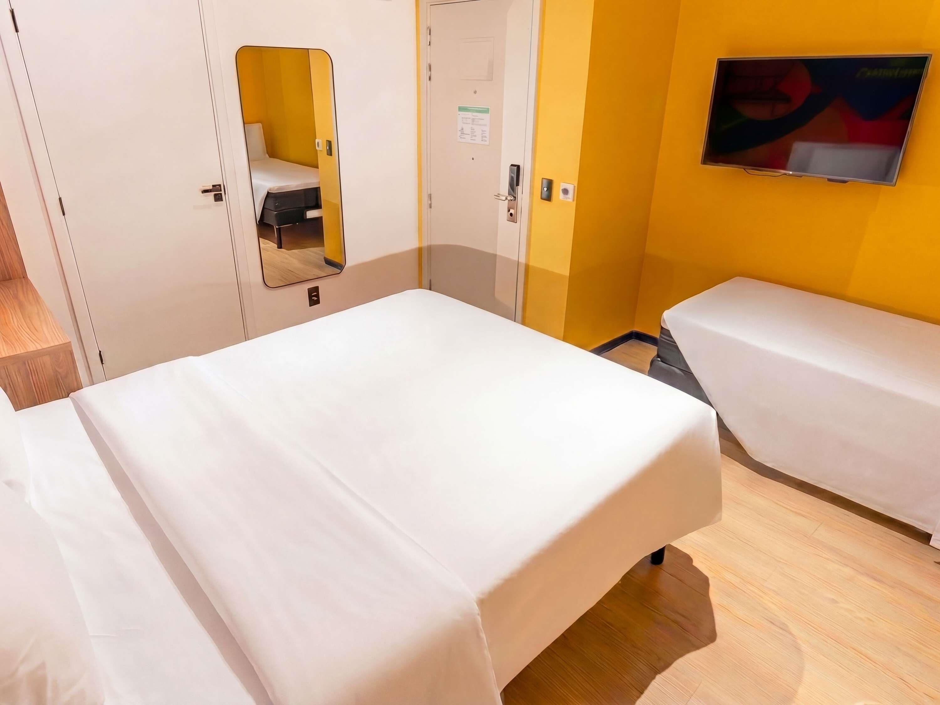 A hotel room with two beds, a mirror, a television, and a door with a small window.