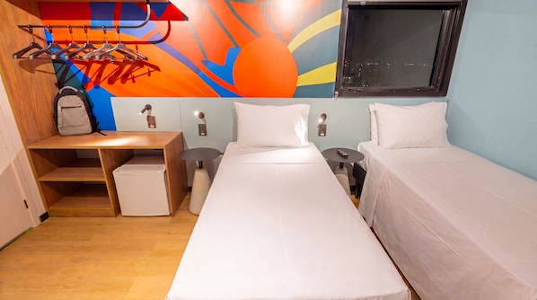 Une chambre d’hôtel avec deux lits, un bureau avec une chaise, une petite table, un porte-manteau et une fenêtre.