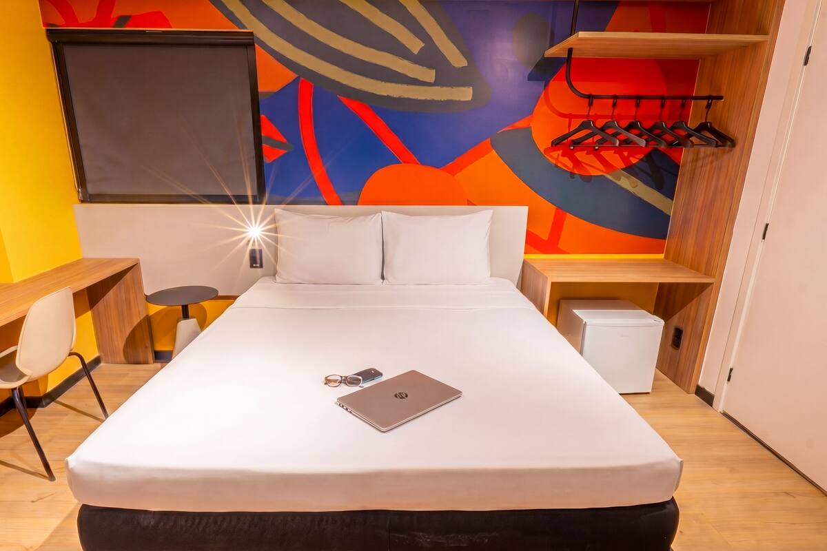 A modern hotel room with a bed, desk, chair, and a wall with abstract art.