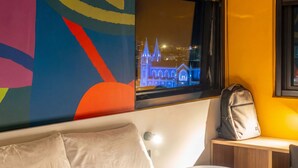 Ein Zimmer mit einem Bett, einem Fenster mit Blick auf ein beleuchtetes Gebäude, einer Wand mit farbenfroher Kunst und einem Rucksack auf einer Bank.