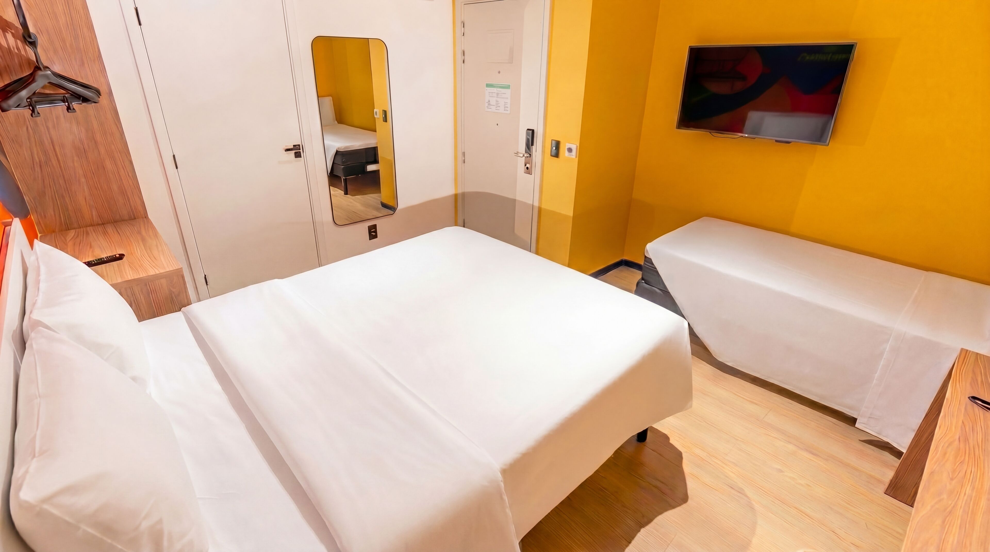 A hotel room with two beds, a wardrobe, a mirror, a television, and a wooden floor.