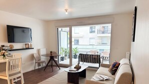 En moderne stue med sofa, sofabord og fjernsyn. Der er en balkon med krukker og planter samt udsigt til bygninger.