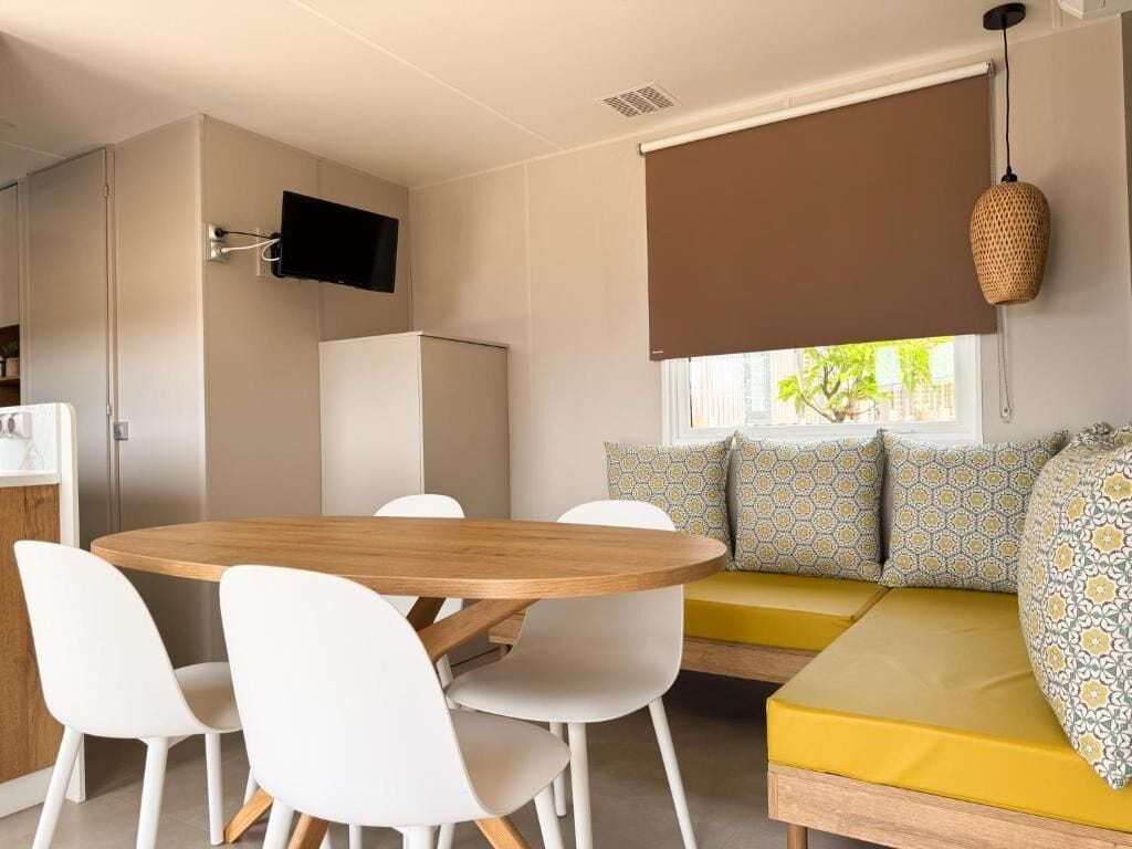 Een knus woongedeelte met een ronde houten tafel, witte stoelen, een bank met decoratieve kussens, een aan de muur bevestigde televisie en een raam met bruin vouwgordijn.