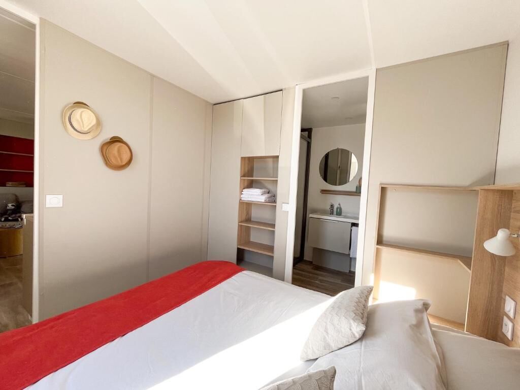 Une chambre à coucher avec un lit, une couverture rouge, une armoire, un miroir et une étagère avec des chapeaux.
