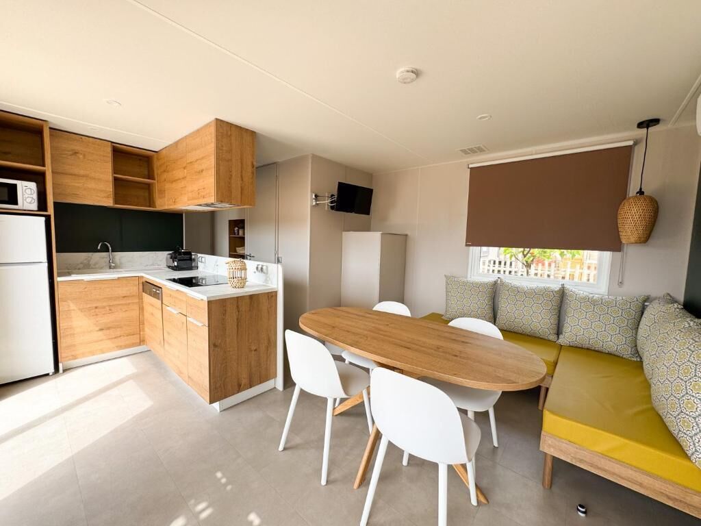 A compact kitchen with wooden cabinets, a dining table with chairs, and a yellow sofa with patterned cushions.