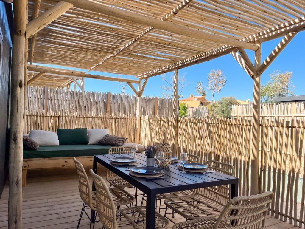 An outdoor patio with a wooden table set for dining, wicker chairs, a cushioned seating area, and a bamboo fence.