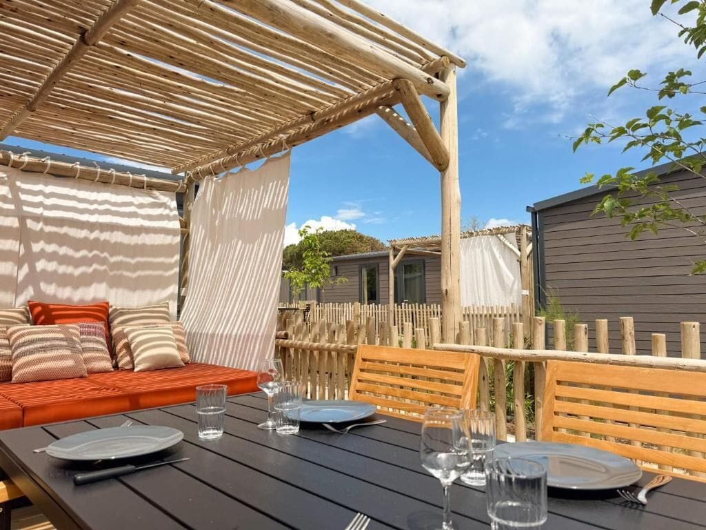 An outdoor dining area with a wooden table set for a meal, a wooden bench, and a covered seating area with cushions.