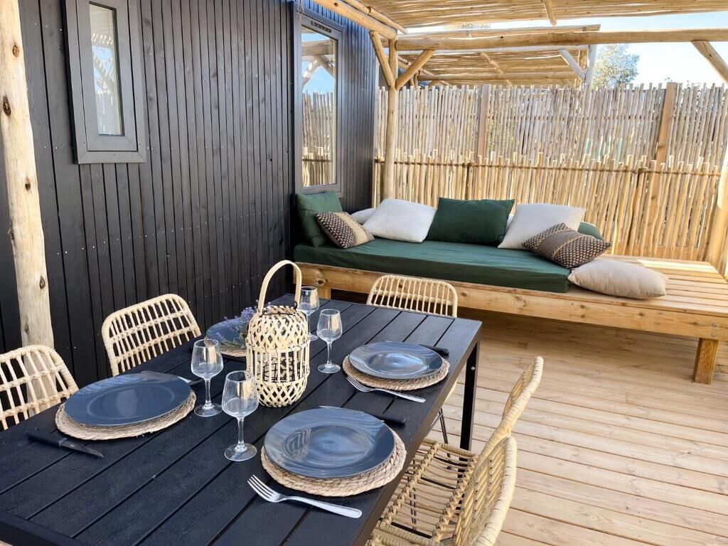 A wooden deck with a dining table set for four, a cushioned bench, wicker chairs, and a wooden fence in the background.