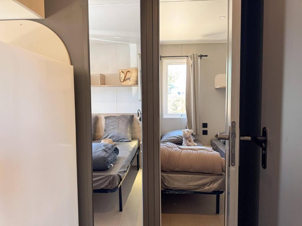 A room with two beds, a window with curtains, a bedside table, and a wall-mounted shelf with a decorative item.