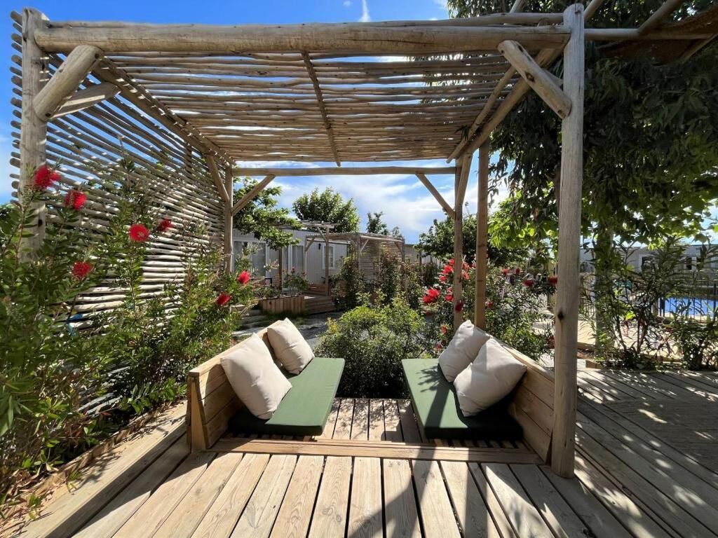 A wooden pergola with a wooden deck, two green cushioned seating areas, and red flowers.
