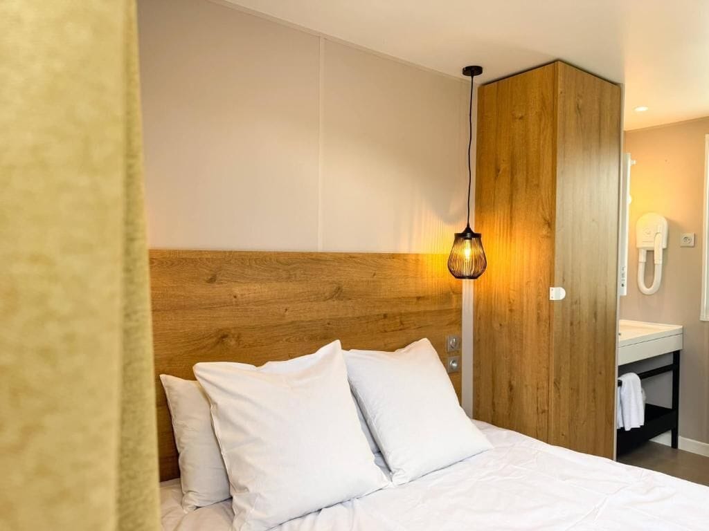 A hotel room with a wooden headboard, a hanging light fixture, a white bed, and a bathroom with a mirror and towel rack.