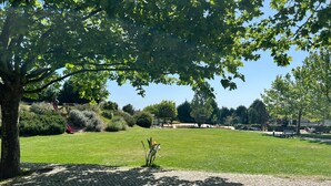 Un área pavimentada con un árbol y césped, un parque infantil con columpios y un banco.